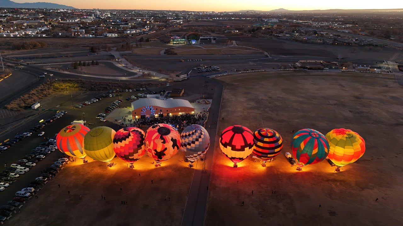 The Night Glow from the air: nine hot air balloons lit in a row on the field, the crowd and the tent beside them